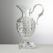 A transparent acrylic pitcher with a geometric pattern and a silver handle and base, placed on a grey surface.