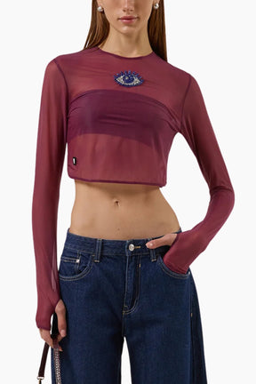 Person wearing a burgundy long-sleeve crop top with an eye design and blue jeans on a white background