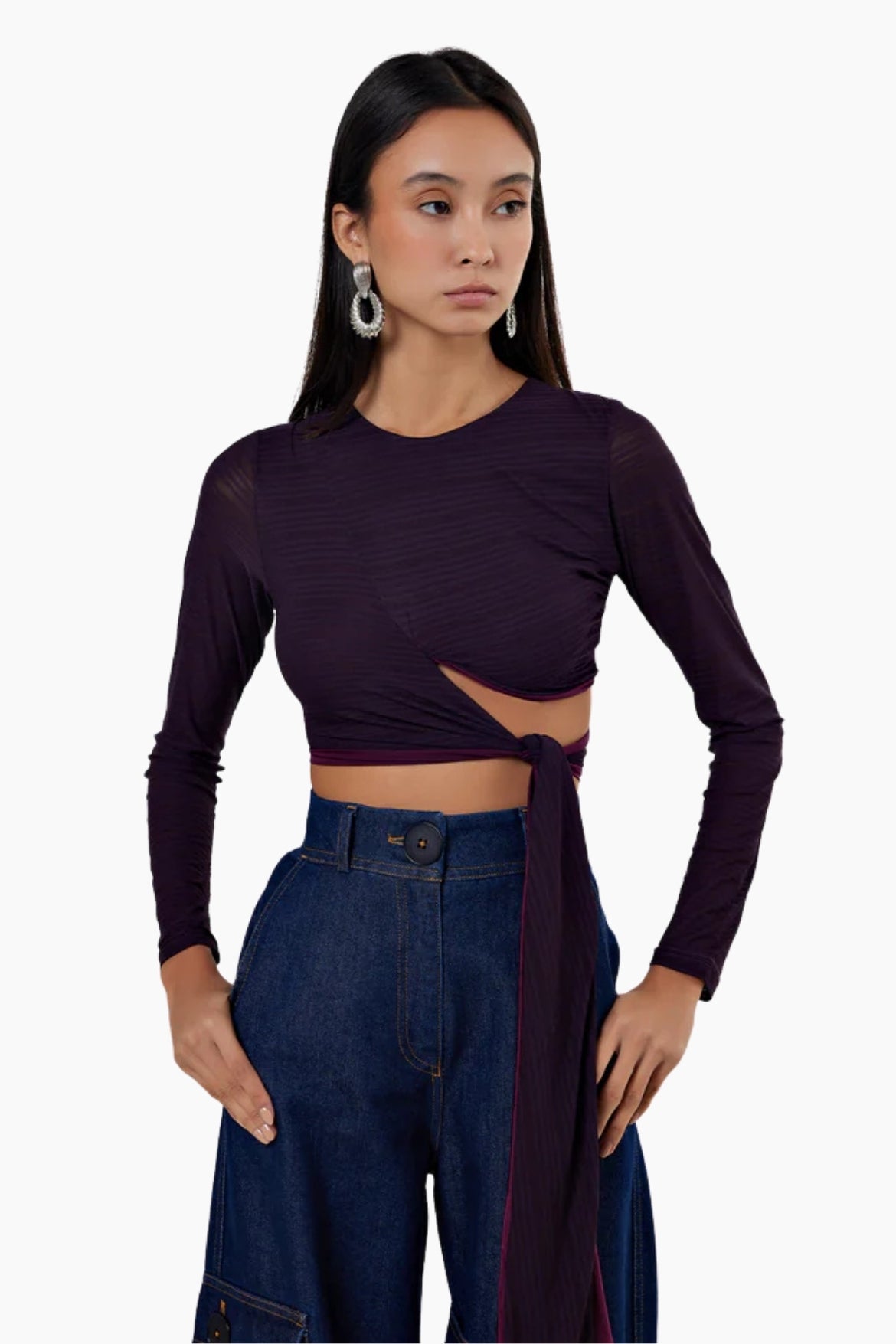 Woman wearing a dark purple long-sleeve top with cut-out design and high-waisted blue jeans on a white background