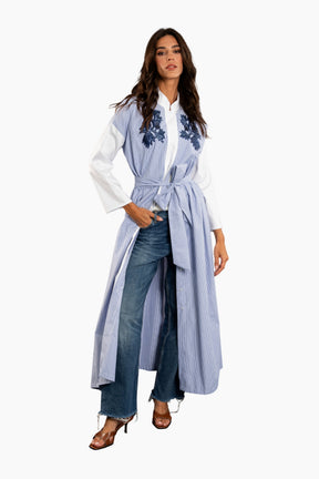 Woman wearing a long light blue embroidered jacket over a white blouse and jeans on a white background