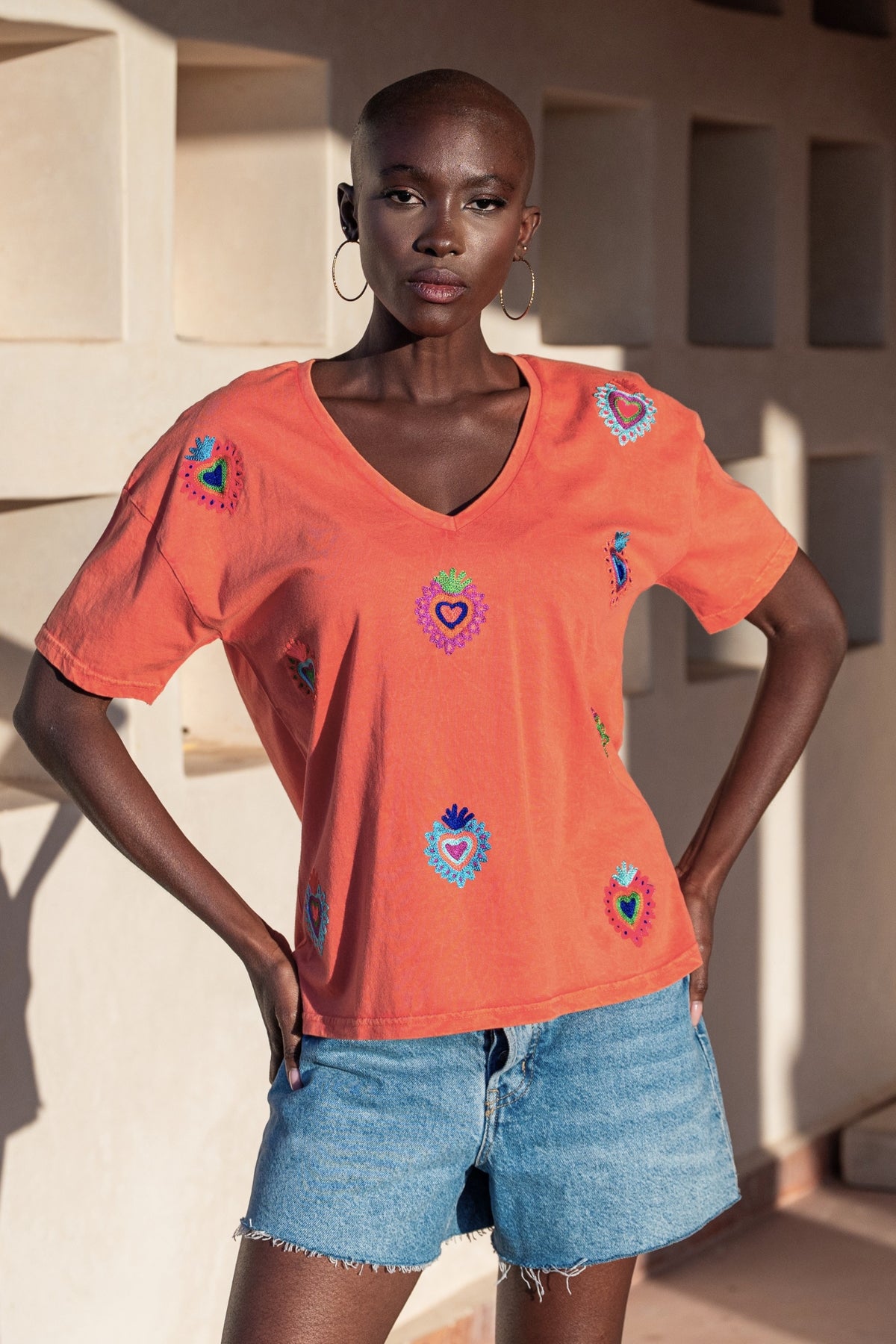 Woman wearing an orange t-shirt with colorful heart designs and denim shorts against a neutral background