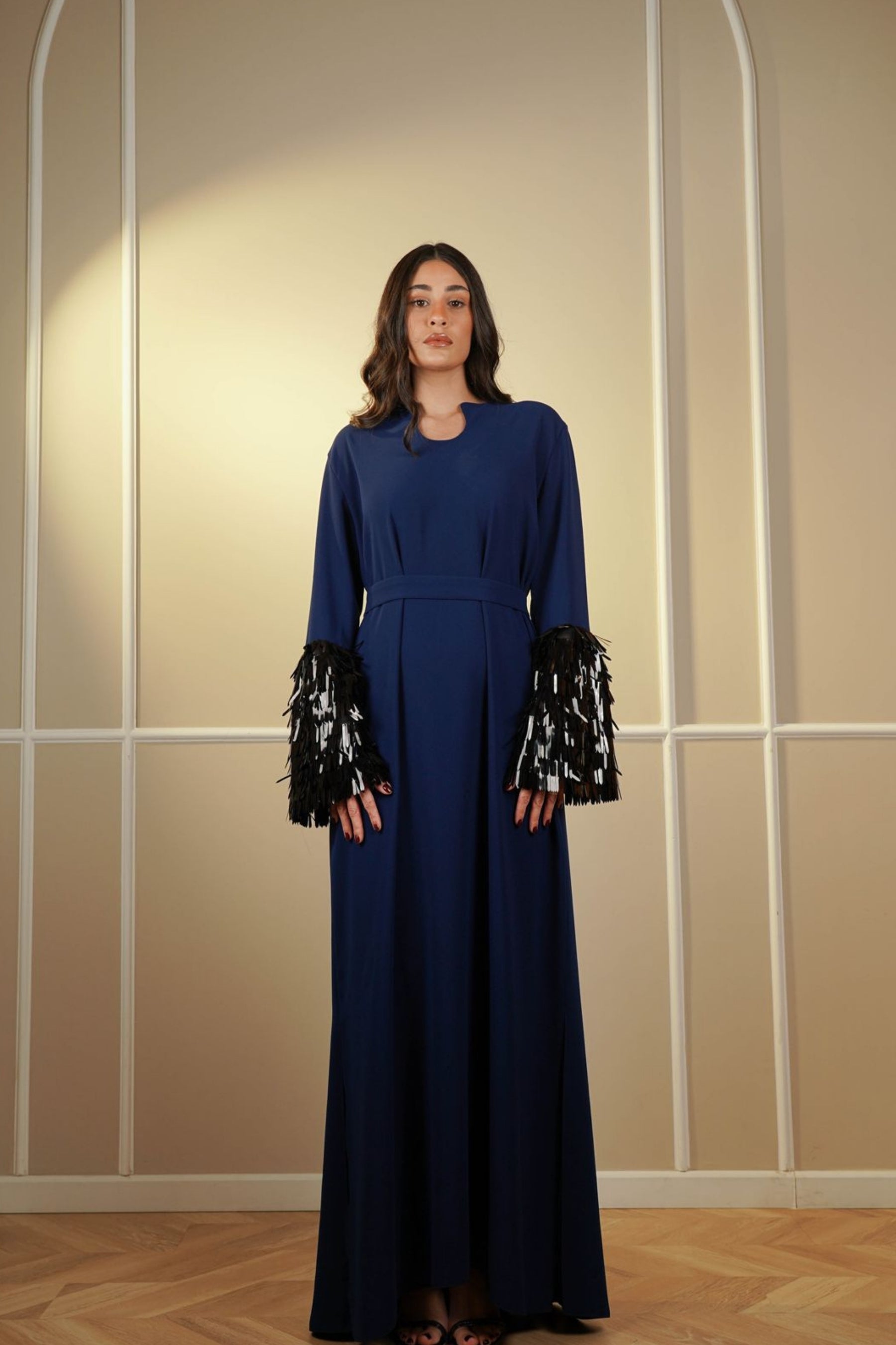 Woman wearing a long blue dress with black and white patterned sleeves against a beige wall.