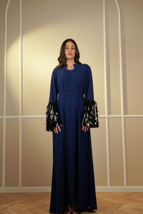 Woman wearing a long blue dress with black and white patterned sleeves against a beige wall.