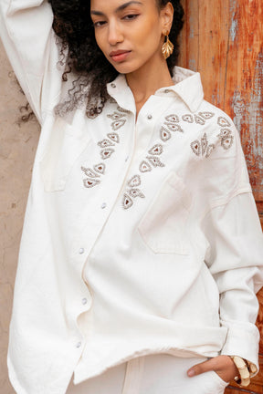 Woman wearing a white embroidered blouse against a wooden wall.