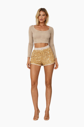 Woman wearing a beige long-sleeve top and gold sequin shorts on a white background