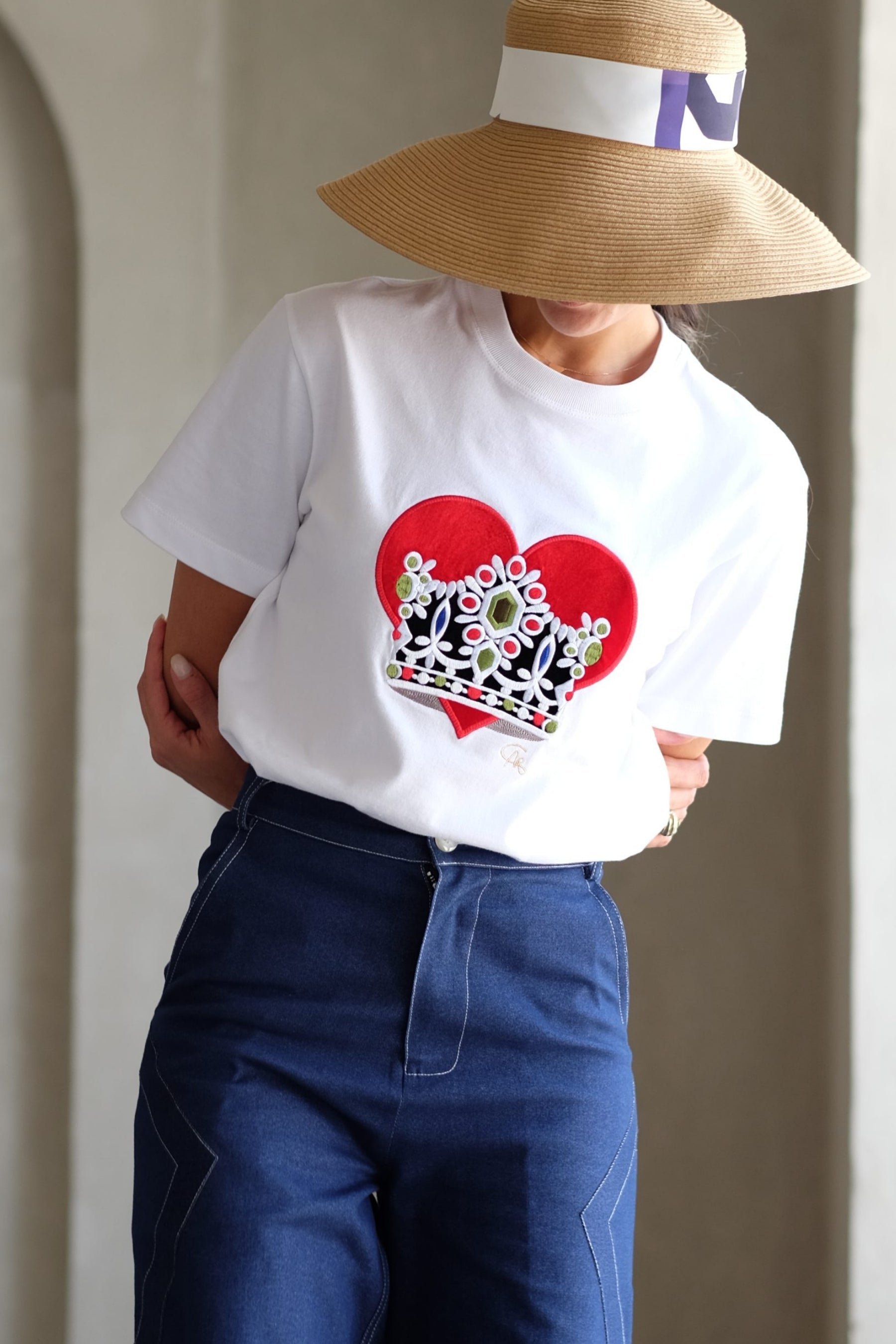 Person wearing a white t-shirt with a red heart and crown design, blue jeans, and a wide-brimmed hat.