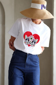 Person wearing a white t-shirt with a red heart and crown design, blue jeans, and a wide-brimmed hat.